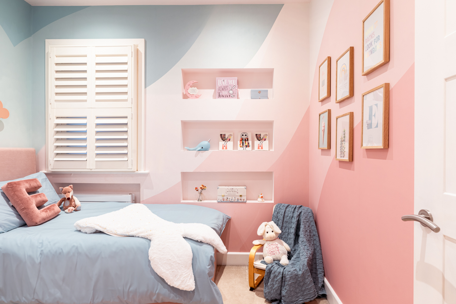 Little girl's bedroom feature a pink and teal mural wallpaper, storage bed, sitting chair and recessed shelving.